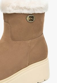 Bottine en suède synthétique marron avec une doublure en fausse fourrure blanche en haut, présentant un accent en métal doré et une semelle compensée de couleur crème.