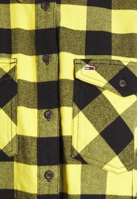 Camicia flanella a quadri gialla e nera con due tasche sul petto, bottoni marroni e un piccolo etichetta con logo. Tessuto testurizzato con un motivo a scacchi.