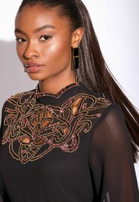 Černé oděvy s propracovanými, texturovanými korálky v zlatých a burgundských vzorech kolem výstřihu. Modelka má na sobě obdélníkové černé náušnice.