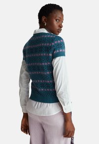 Pletený svetřík bez rukávů v tyrkysové barvě s růžovými geometrickými vzory, doplněný bílou košilí s límečkem. Žebrovaný lem a krátké rukávy.