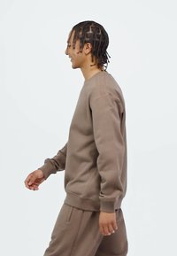 Man with braided hair wearing a brown sweatshirt and matching pants walking against a plain white background, smiling.