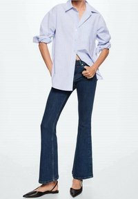 Camisa de rayas azul claro y blanca de corte oversized, jeans acampanados azul oscuro y slippers negras de punta. La camisa tiene mangas enrolladas; los jeans tienen un ajuste clásico.