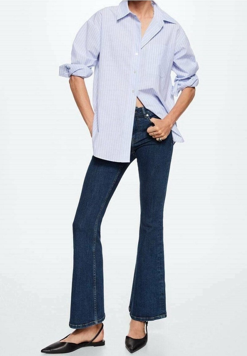 Camisa de rayas azul claro y blanca de corte oversized, jeans acampanados azul oscuro y slippers negras de punta. La camisa tiene mangas enrolladas; los jeans tienen un ajuste clásico.