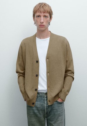 Brown cardigan with a textured knit, featuring a v-neck, five buttons, and ribbed cuffs. Worn over a white shirt and paired with denim jeans.