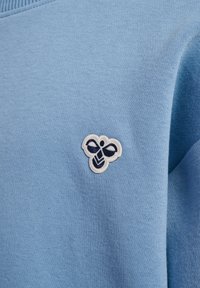 Lichtblauwe sweater met een getextureerde brei, met een wit en donkerblauw geborduurd bijenlogo op de linkerborst.