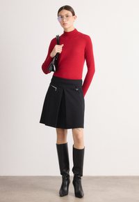 Czerwony sweter z golfem, czarna spódnica plisowana z zasuwanym kieszeniem po boku i wysokie czarne kozaki. Akcesoria to mała czarna torba.