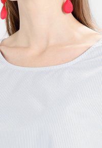 Haut à rayures bleu clair avec un col arrondi. Met en avant des boucles d'oreilles rouges en forme de goutte. Texture de tissu lisse, motif minimaliste.