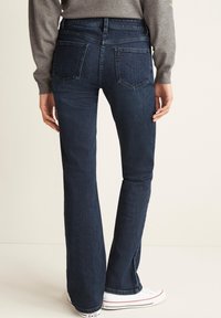Jean évasé en denim bleu foncé avec une taille mi-haute, deux poches arrière et une texture douce, associé à des baskets blanches en toile.