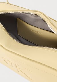 Sárga bőrtáska, sima felülettel, ezüst cipzárral és szürke belső térrel. Jellemzői között szerepel a dombornyomott "CK" logó és egy kártyahely.