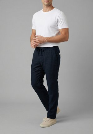 Uomo che indossa una t-shirt bianca a maniche corte, pantaloni blu scuro con coulisse e sneakers beige casual, in piedi con le mani giunte davanti.