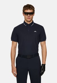 Marinblå polo skjorta med vit krage och logotyp, i kombination med svarta byxor och handskar, bärs av en manlig figur.