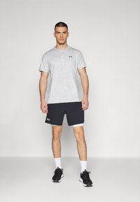Camiseta corta de manga gris con una textura sutil y un logo verde, combinada con pantalones cortos deportivos negros y zapatillas de correr negras. Calcetines blancos visibles.