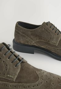 Ciemnooliwkowe zamszowe buty z perforowanym wzorem, okrągłym noskiem oraz czarną gumową podeszwą. Posiadają pasujące sznurówki i kontrastujące akcenty w szyciu.