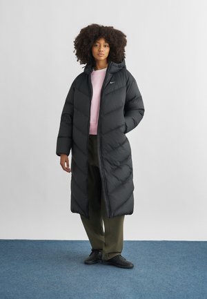 Žena s kudrnatými vlasy, oblečená v dlouhém černém péřovém kabátu, světle růžovém svetru, olivových kalhotách a černých teniskách stojící na modrém koberci.