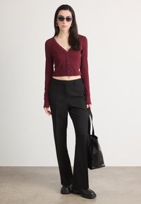 Top scurt de culoare vișinie cu nasturi și mâneci lungi, asortat cu pantaloni negri evazați. Modelul poartă o geantă de umăr neagră și ochelari de soare închiși la culoare.