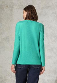 Maglione in turchese con maniche lunghe, dettaglio testurizzato sulle spalle e una vestibilità rilassata. Abbinato a jeans blu scuro.