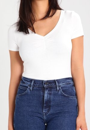 Haut blanc à manches courtes avec col en V et détail froncé au niveau de la poitrine, associé à un jean en denim bleu foncé taille haute avec fermeture à boutons.