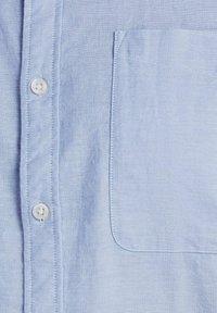 Camisa de botones azul claro hecha de una tela suave, con un bolsillo en el pecho. Presenta botones blancos y costuras cuidadas a lo largo de los bordes.