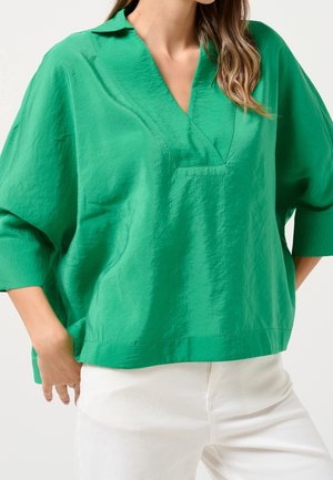 Femme portant un chemisier vert vif à col en V avec des manches trois-quarts et un pantalon blanc, main reposant près de la poche.