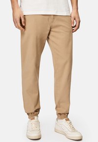 Beige, leichte Jogginghosen mit elastischen Bündchen und seitlichen Taschen, kombiniert mit weißen Sneakern. Glattes Baumwollmaterial, lockere Passform.