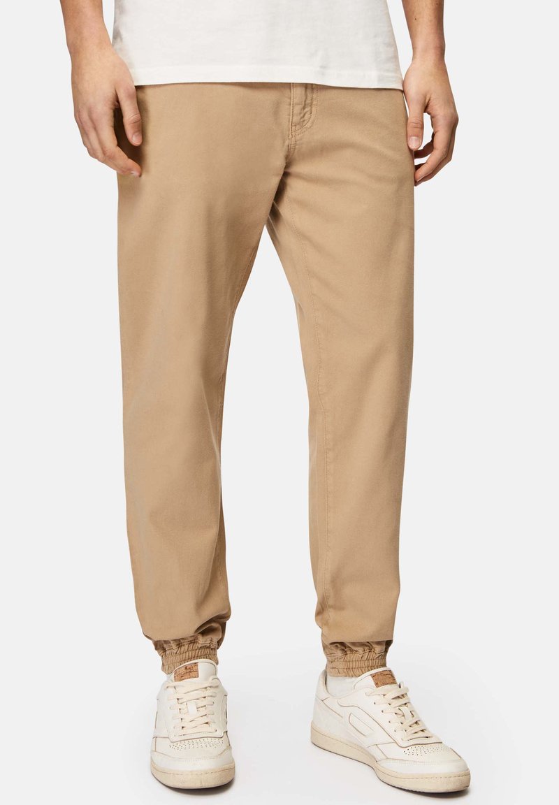 Beige, leichte Jogginghosen mit elastischen Bündchen und seitlichen Taschen, kombiniert mit weißen Sneakern. Glattes Baumwollmaterial, lockere Passform.