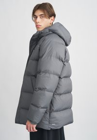 Casaco puffer cinza com capuz, apresentando um design acolchoado, acabamento mate e punhos elásticos. Logótipo exibido no lado inferior esquerdo.