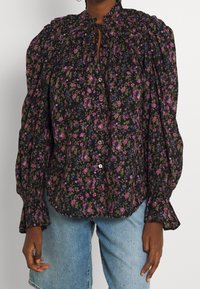 Svart blommig blus med puffärmar, prydd med rosa och blå blommor, hög hals, knäppning och draperade detaljer vid ärmsluten.