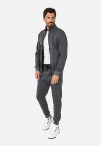 Tuta grigia con zip e colletto alto, indossata con una T-shirt bianca sotto e pantaloni neri con cordino. Completata da sneaker bianche.