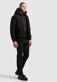 Czarna pikowana kurtka z wysokim kołnierzem, noszona na pikowanej kamizelce, w zestawieniu z ciemnymi dżinsami i czarnymi, sznurowanymi butami. Gładka faktura i smukły design.