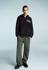 Fekete bordázott cipzáras kapucnis pulóver elülső zsebekkel és fehér grafikai designnal. Laza szürke nadrággal és fekete cipővel párosítva.