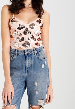 Nainen pukeutunut hihattomaan, suurilla paljeteilla koristeltuun vaaleanpunaiseen lyhytpaitaan ja korkeavyötäröisiin, kuluneisiin sinisiin farkkuihin yksivärisen taustan edessä.