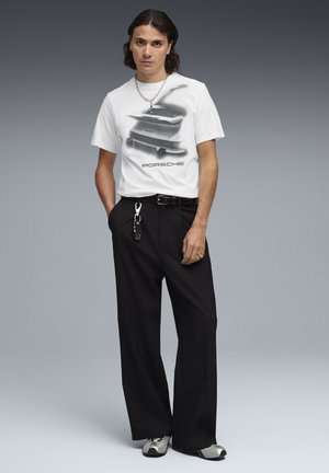T-shirt blanc avec un motif graphique noir d'une Porsche, accompagné d'un pantalon large noir. Les accessoires comprennent un porte-clés et des chaussures argentées.