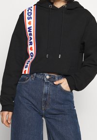 Sweat à capuche court noir avec bandes contrastantes en rouge, blanc et bleu, portant les textes "GCDS" et "WEAR". Cordon réglable avec embouts métalliques.