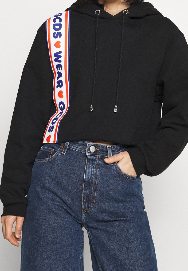 Sweat à capuche court noir avec bandes contrastantes en rouge, blanc et bleu, portant les textes "GCDS" et "WEAR". Cordon réglable avec embouts métalliques.