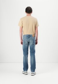 Jean en denim bleu avec une coupe droite, poches arrière standards et une étiquette marron à la taille, associé à un t-shirt beige à manches courtes.