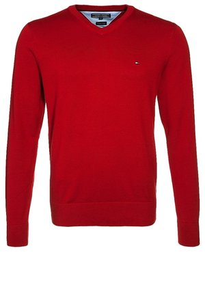 Mężczyzna ubrany w czerwony sweter na szarej koszuli, wyblakłe szare dżinsy i ciemnobrązowe buty, trzymający brązową skórzaną aktówkę w lewej ręce.