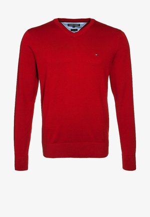 Mann trägt einen roten Pullover über einem grauen Hemd, ausgebleichte graue Jeans und dunkelbraune Stiefel, hält eine braune Ledertasche in der linken Hand.