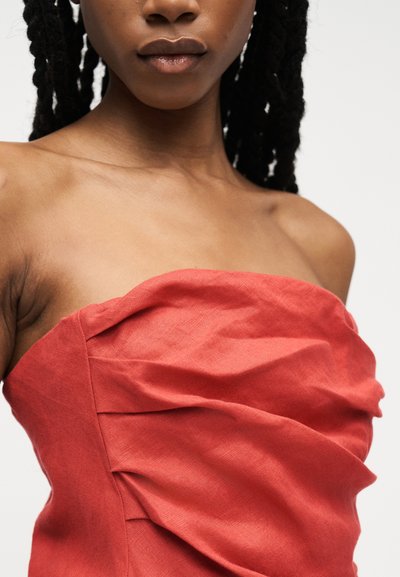 Fiatal fekete nő fonott hajjal, piros, pánt nélküli, redőzött anyagú ruhában, egyszerű háttér előtt.