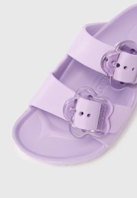 Lilla gummisandal med to brede stropper og gennemsigtige blomsterformede spænder. Glat tekstur med en formet fodseng.