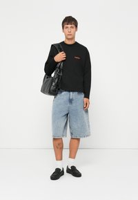 Black long-sleeve shirt with red logo, blue denim shorts, white socks, and black slip-on shoes. A black shoulder bag completes the outfit.