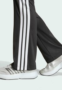 Crne atletske hlače s tri bijele pruge sa strane, u kombinaciji sa srebrnim tenisicama za trčanje koje imaju mrežaste i teksturirane akcentne detalje.