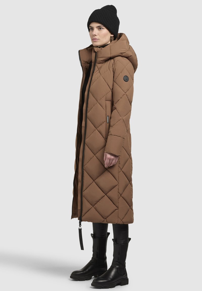 Langer, brauner, gesteppter Daunenmantel mit Kapuze, seitlichen Taschen und einem schwarzen Logo-Patch. Getragen mit schwarzen Leggings und hohen, derben Stiefeln.