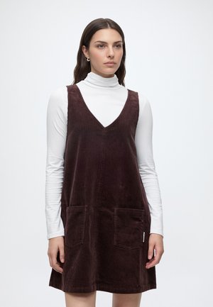 Vrouw met een witte coltrui met lange mouwen onder een mouwloze bruine corduroy jurk met zakken aan de voorkant, staand tegen een effen achtergrond.