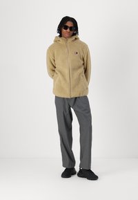 Beige Fleece-Zip-Hoodie mit Kapuze, ausgestattet mit einem kleinen Logo-Patch. Kombiniert mit grauen Hosen und schwarzen Sneakers. Glatte Textur des Stoffes.