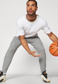 Hvit kortermet skjorte med lilla og gul krage, grå tapered joggebukser og svarte og kremfargede joggesko. Holder en oransje basketball.
