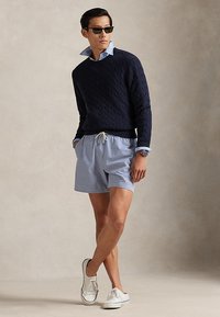 Hombre con gafas de sol, suéter azul marino de punto trenzado sobre camisa azul con cuello, pantalones cortos a rayas y zapatillas blancas sin cordones, de pie frente a un fondo liso.