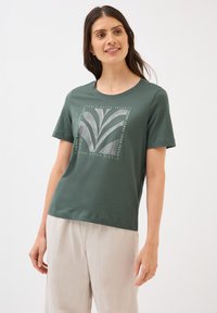 Kvinna med långt brunt hår ler medan hon bär en grön t-shirt med en grafisk design, i kombination med ljusa byxor.