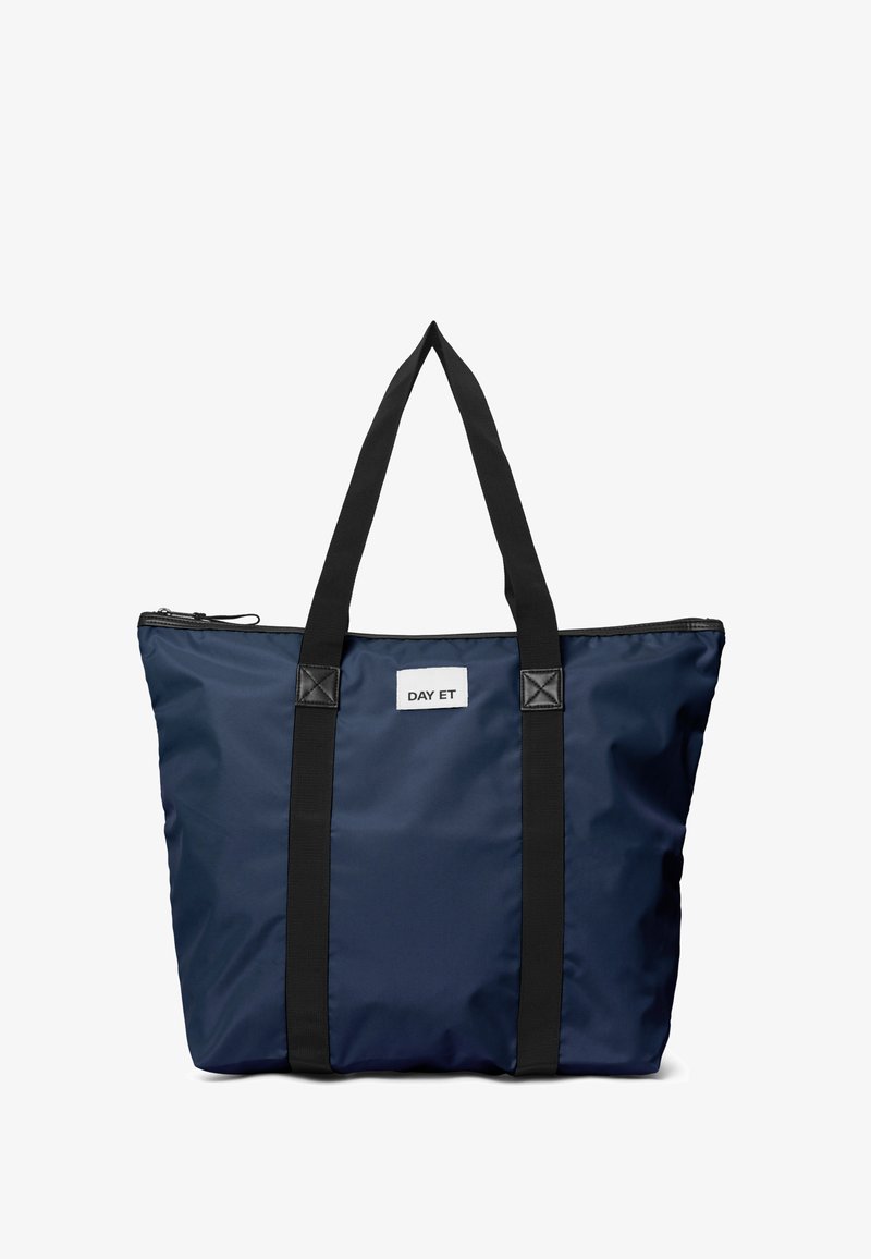 Marineblauwe tas met zwarte banden en rits, gemarkeerd met "DAY ET", ontworpen voor dagelijks gebruik, met een moderne, eenvoudige esthetiek.