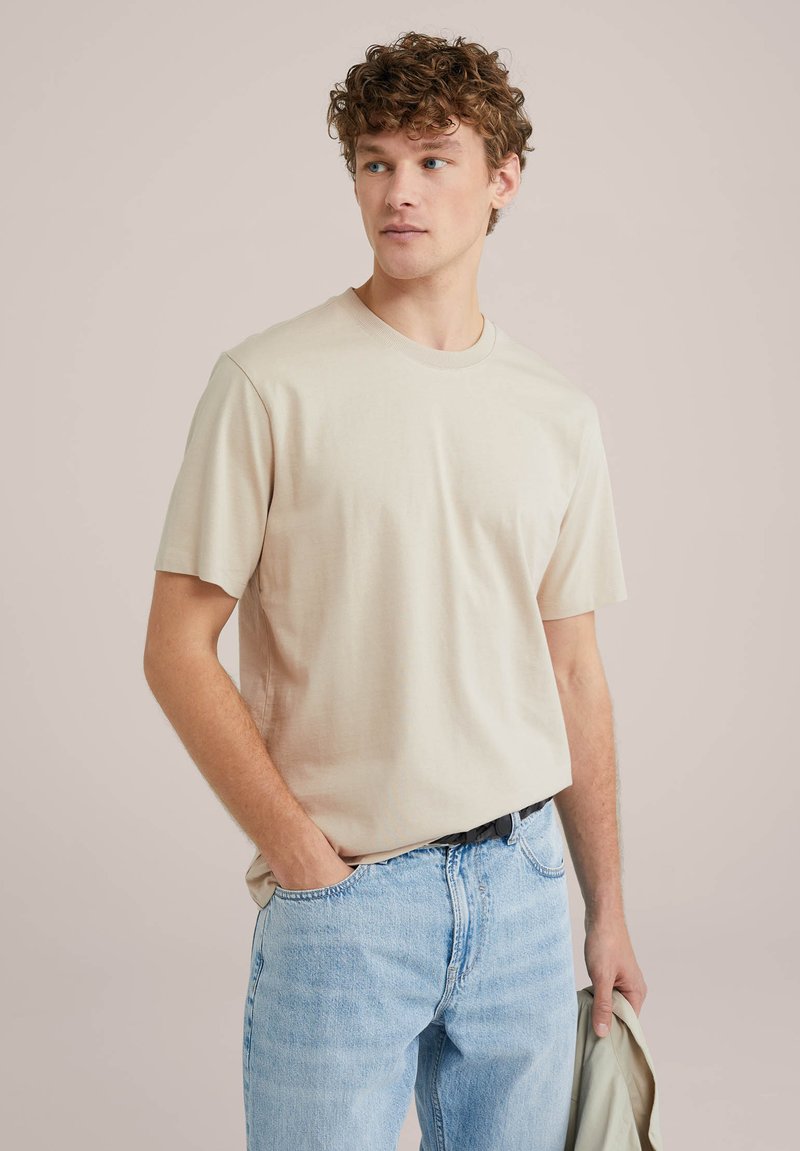 Beige tričko s krátkým rukávem z hladkého materiálu s kulatým výstřihem, spojené se světle modrými džíny, drženo volně jednou rukou.