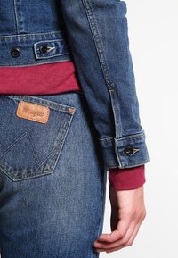 Denim takki sinisellä kankaalla, jossa on mustat napit ja marooni villapaita alla. Farkut, joissa on ruskea nahkainen Wrangler-logo ja ompelu yksityiskohtia.